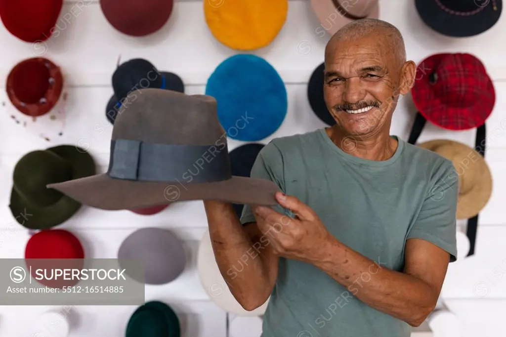 Portrait close up of a senior mixed race man smiling and holding a finished hat in the showroom at a hat factory, with various hats on display in the background