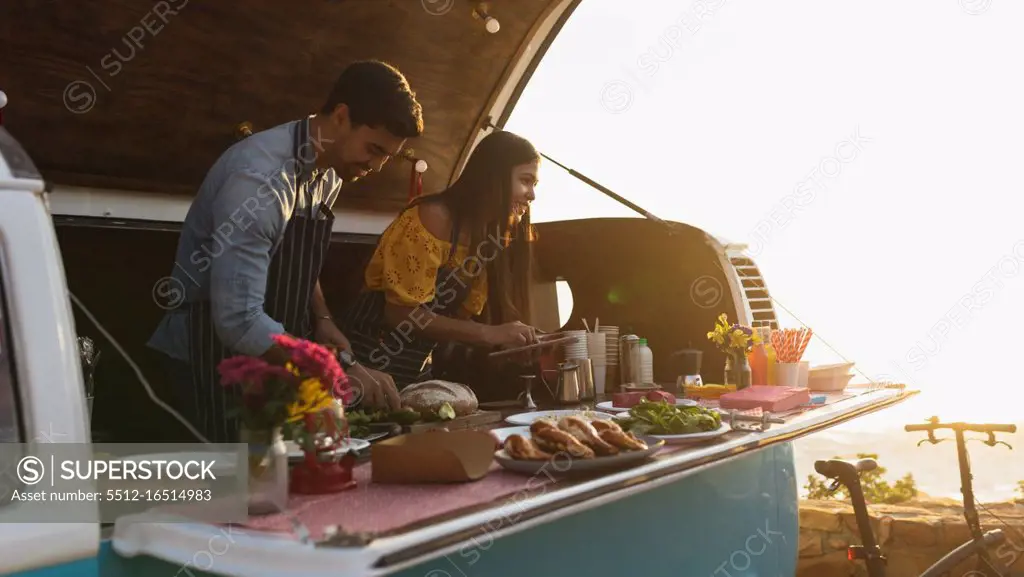 Side view of a young mixed race couple busy preparing and serving food standing in an open top van offering a range of takeaway food for sale