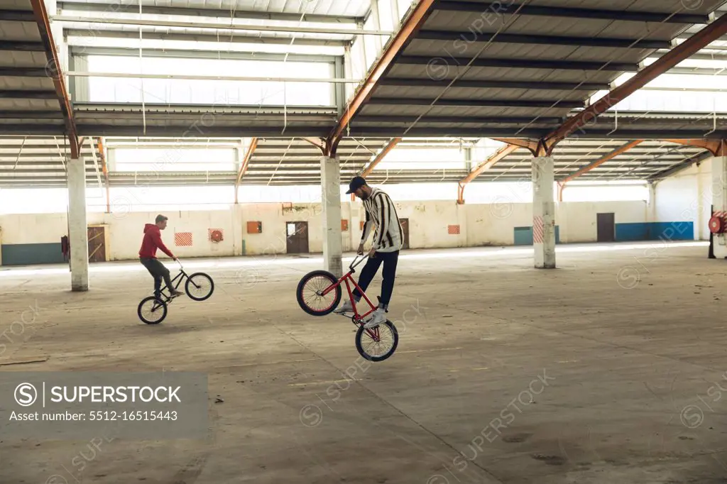 Side view of two young Caucasian men facing opposite directions balancing on the back wheels of their BMX bikes while practicing tricks in an abandoned warehouse, the rider in the foreground is jumping off the ground