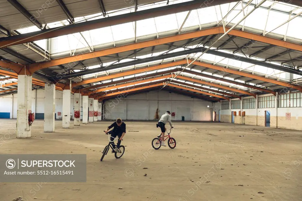 Front view of two young Caucasian men facing opposite directions riding BMX bikes while practicing tricks in an abandoned warehouse