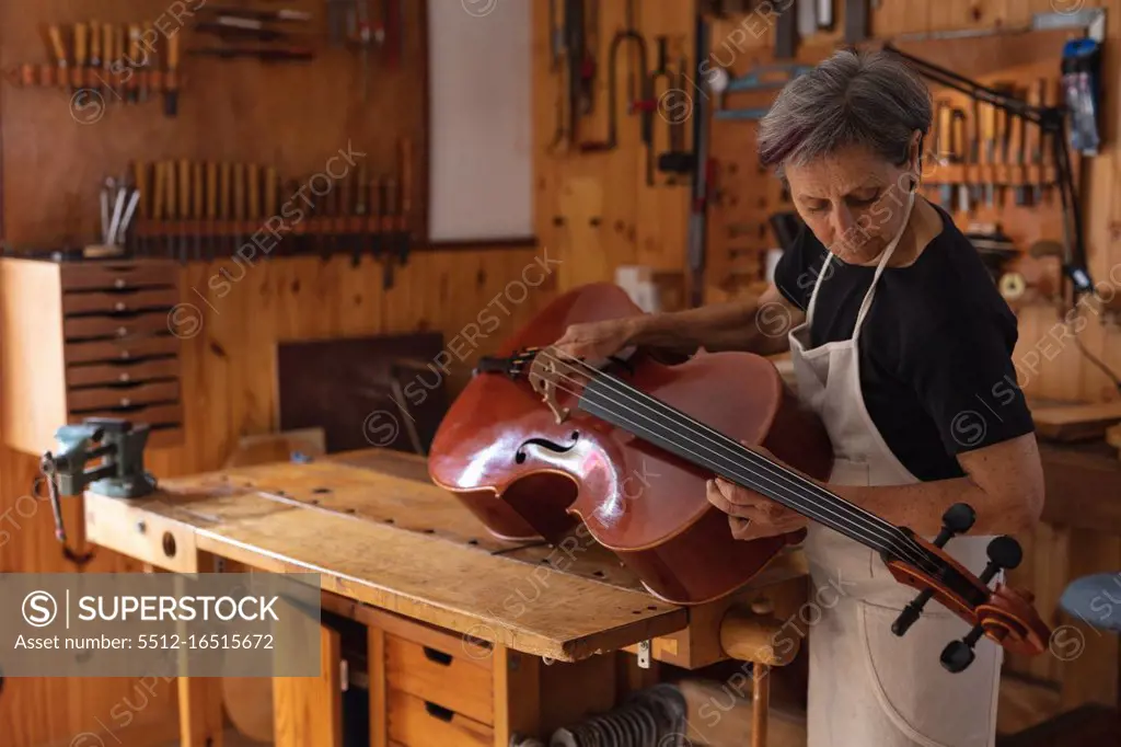 Front view of a senior Caucasian female luthier holding a cello in her workshop with tools hanging up on the wall in the background