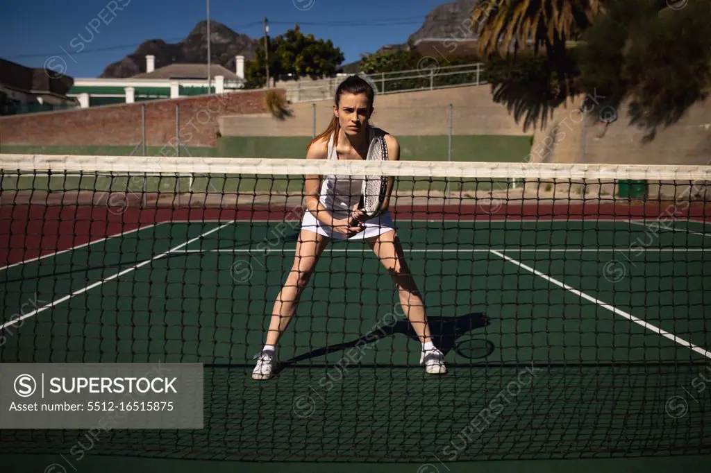 Front view of a young Caucasian woman playing tennis on a sunny day, holding a racket and waiting for the ball, seen through the net