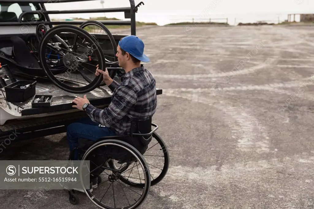 Side view of a young Caucasian man in a wheelchair taking a recumbent bicycle out of the back of his car to assemble it in a car park by the sea