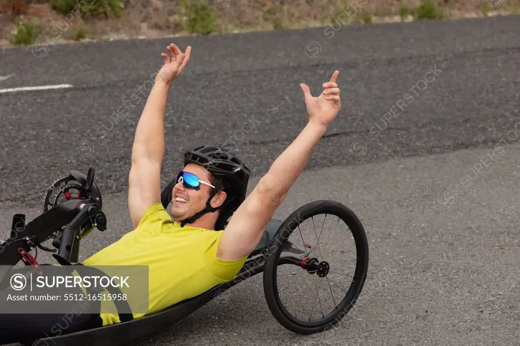 Side view close up of a young Caucasian man in sportswear on a recumbent bicycle cycling on a country road, smiling with his arms in the air