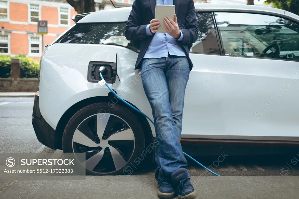 Low section of businessman using digital tablet while charging electric car at charging station