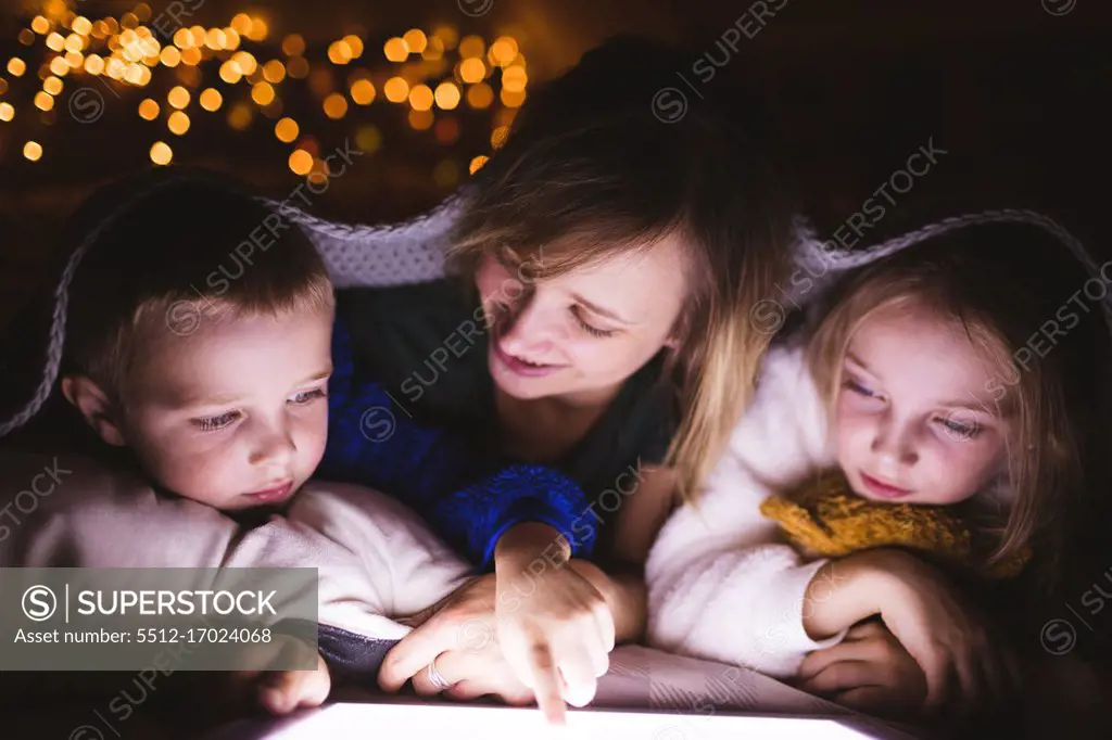 Close-up of mother and kids under the blanket using digital tablet against Christmas lights