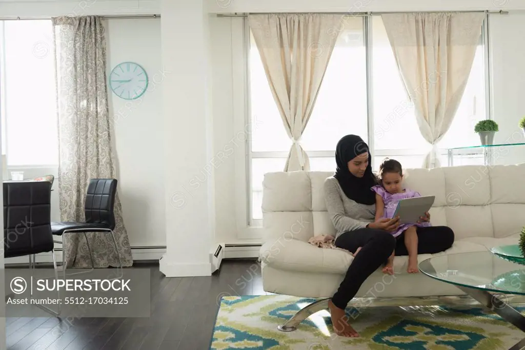 Front view of mixed race mother wearing hijab and daughter using digital tablet in living room at home 