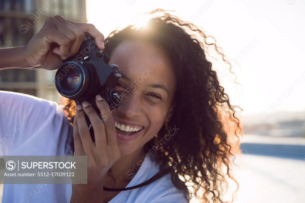 Front view of a young African-American woman smiling while holding a camera and standing on a rooftop with a view of a building and the sunlight. Bright modern gym with fit healthy people working out and training