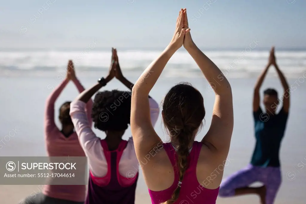 Rear view of a multi-ethnic group of female friends enjoying exercising on a beach on a sunny day, practicing yoga, standing in yoga position.