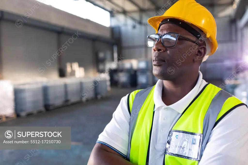Portrait of Thoughtful African-american male staff in hardhat and reflective jacket standing with arms crossed in warehouse. This is a freight transportation and distribution warehouse. Industrial and industrial workers concept