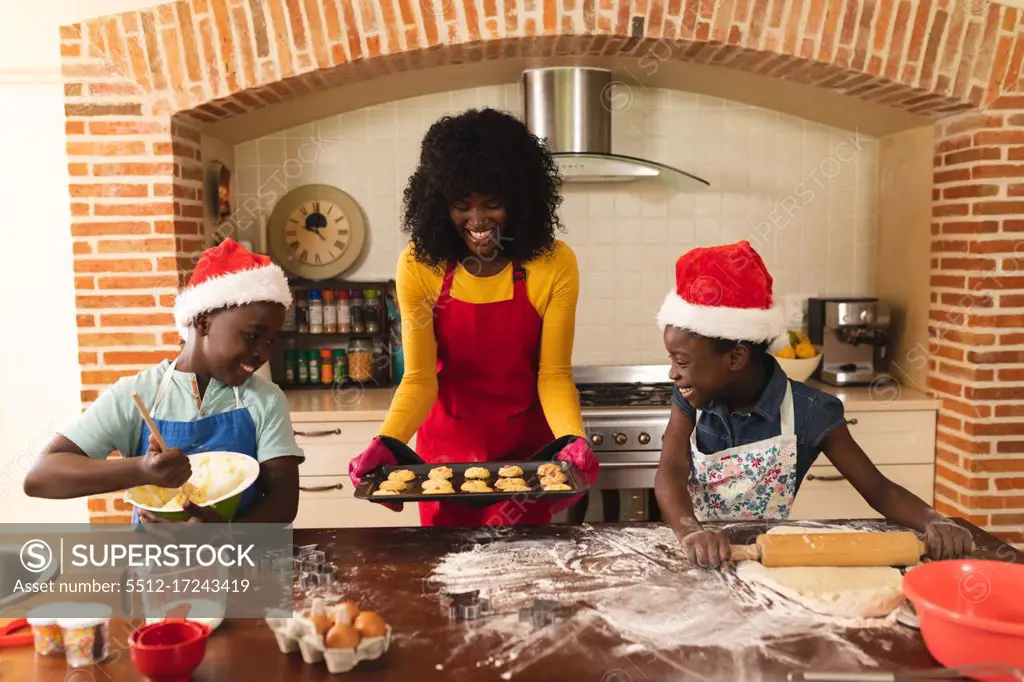 African american mother wearing apron holding baked cookies and daughter and son wearing santa hat baking in the kitchen at home. christmas festivity tradition celebration concept