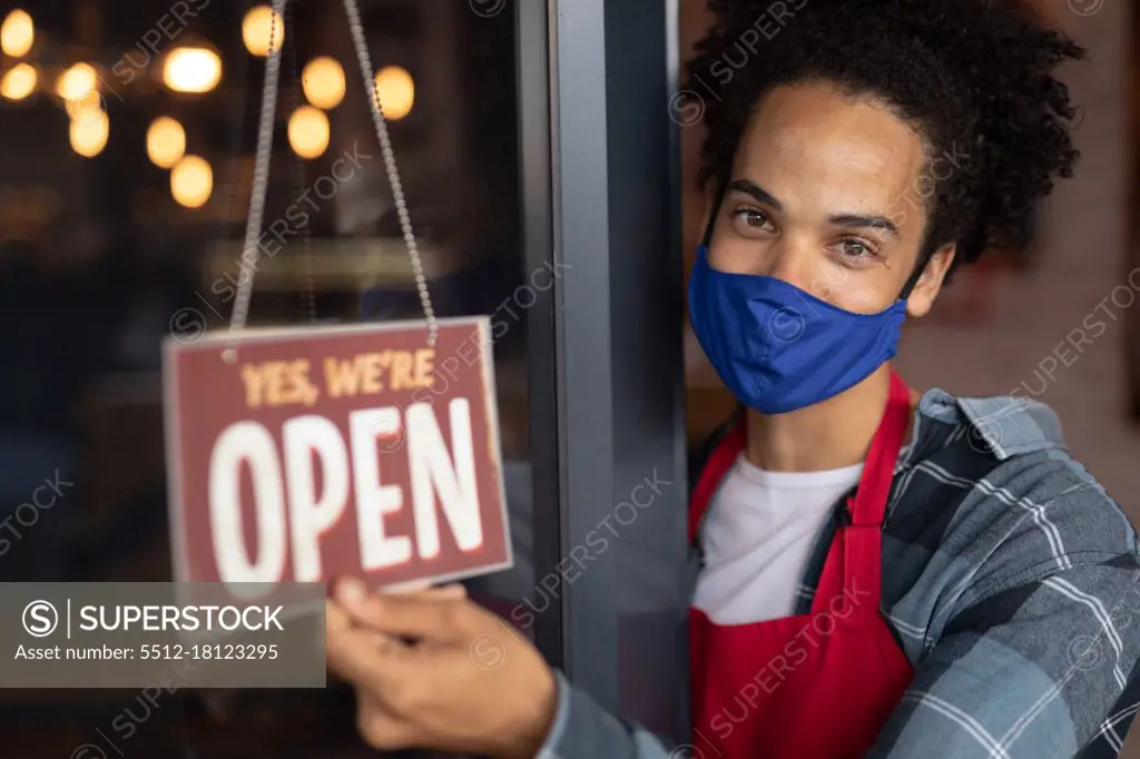 Portrait of mixed race male barista wearing face mask leaning in doorway of cafe holding open sign. independent small business during coronavirus covid 19 pandemic.