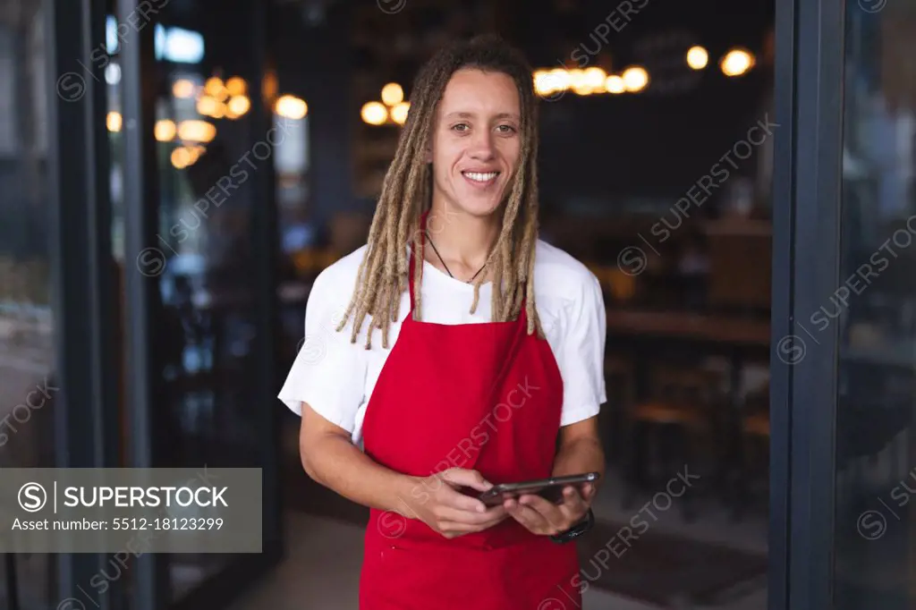 Portrait of happy mixed race male barista with dreadlocks standing in doorway of cafe holding tablet. independent small business in a city.
