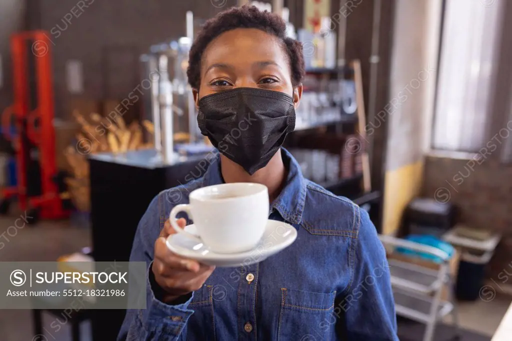 Portrait of african american woman wearing face mask holding a coffee cup at a cafe. health protection and safety during covid-19 pandemic concept