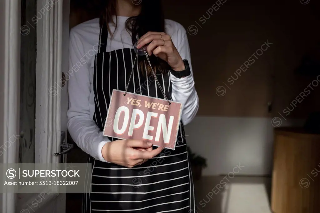 Midsection of caucasian waitress wearing striped apron, standing in doorway, holding open sign. small independent cafe business.