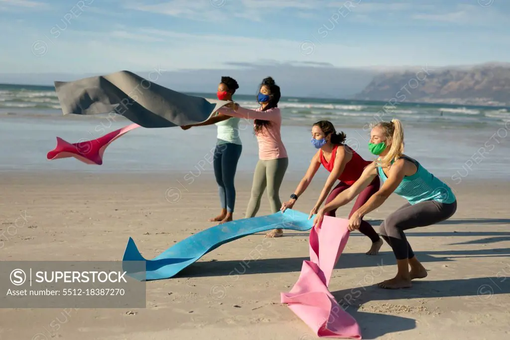 Group of diverse female friends wearing face masks putting down mats, practicing yoga, at the beach. healthy active lifestyle, outdoor fitness and well being during covid 19 pandemic.