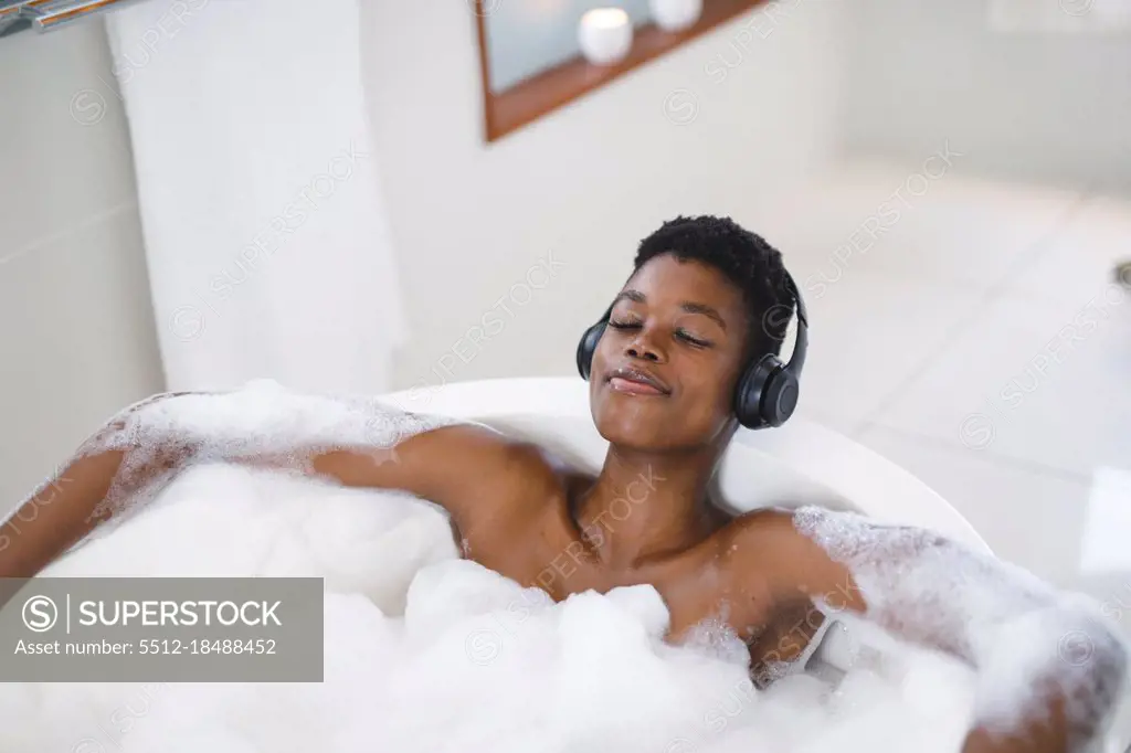 Smiling african american woman in bathroom relaxing in foam bath wearing headphones with eyes closed. domestic lifestyle, enjoying self care leisure time at home.