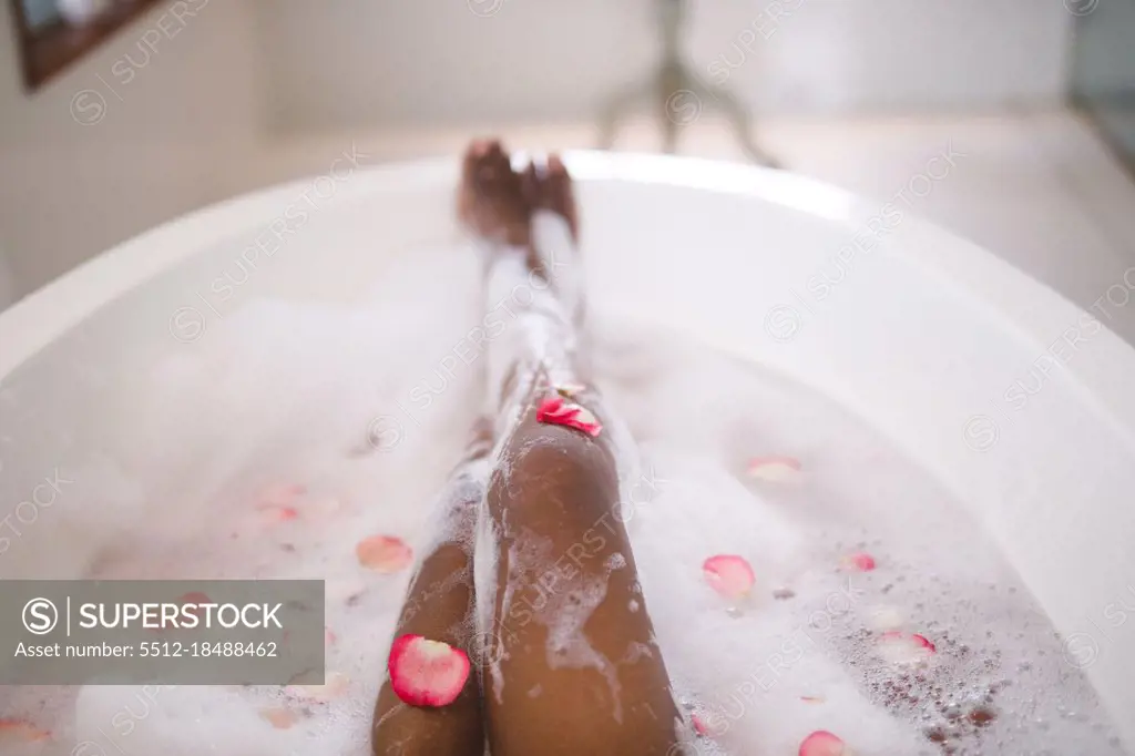 Low section of african american woman relaxing in pampering foam bath with rose petals. domestic lifestyle, enjoying self care leisure time at home.