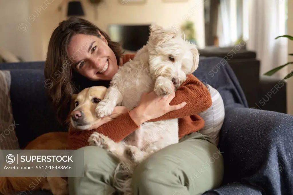 Portrait of smiling caucasian woman in living room sitting on sofa embracing her pet dogs. domestic lifestyle, enjoying leisure time at home.