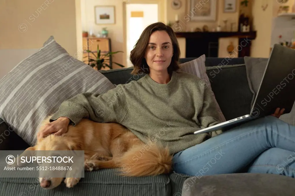 Portrait of smiling caucasian woman in living room, sitting on sofa with her pet dog, using laptop. domestic lifestyle, enjoying leisure time at home.