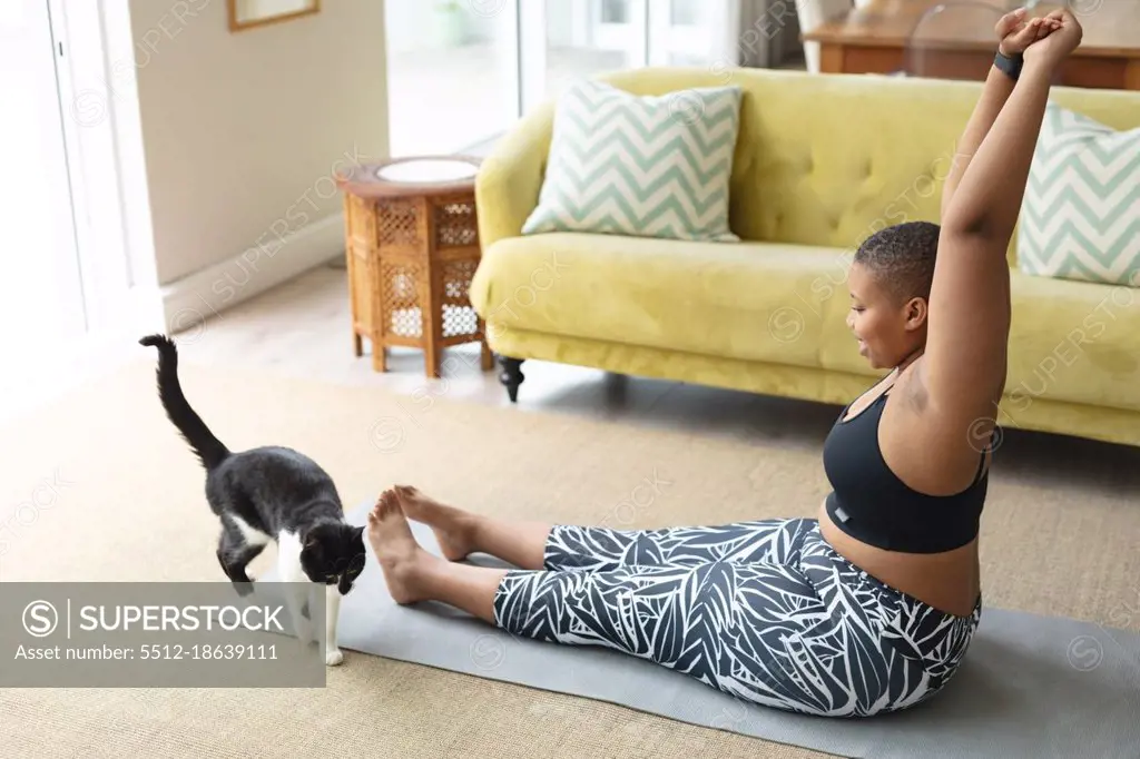 Happy african american plus size woman practicing yoga on mat at home with cat. fitness and healthy, active lifestyle.
