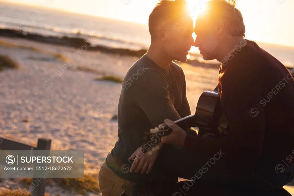 Happy caucasian gay male couple playing guitar and kissing at sundown by the sea. summer road trip and holiday in nature.