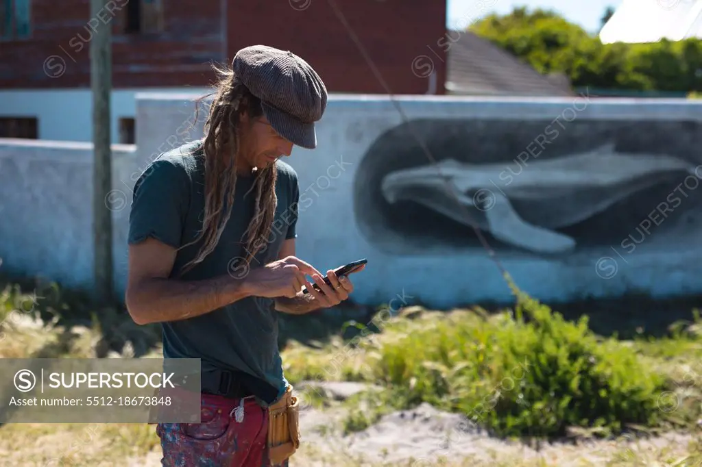 Male mural artist using smart phone while standing near whale painting on wall during sunny day. technology, street art and skill.