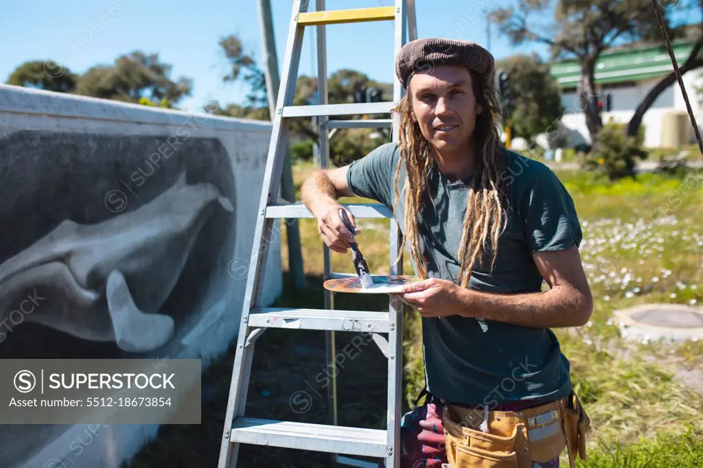 Portrait of male artist holding paintbrush and palette leaning on ladder by whale mural on wall. street art and skill.