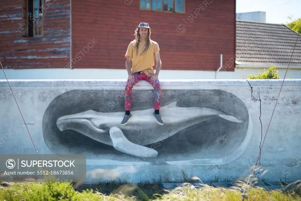Portrait of male artist sitting on wall with whale mural painting against house. street art and skill.