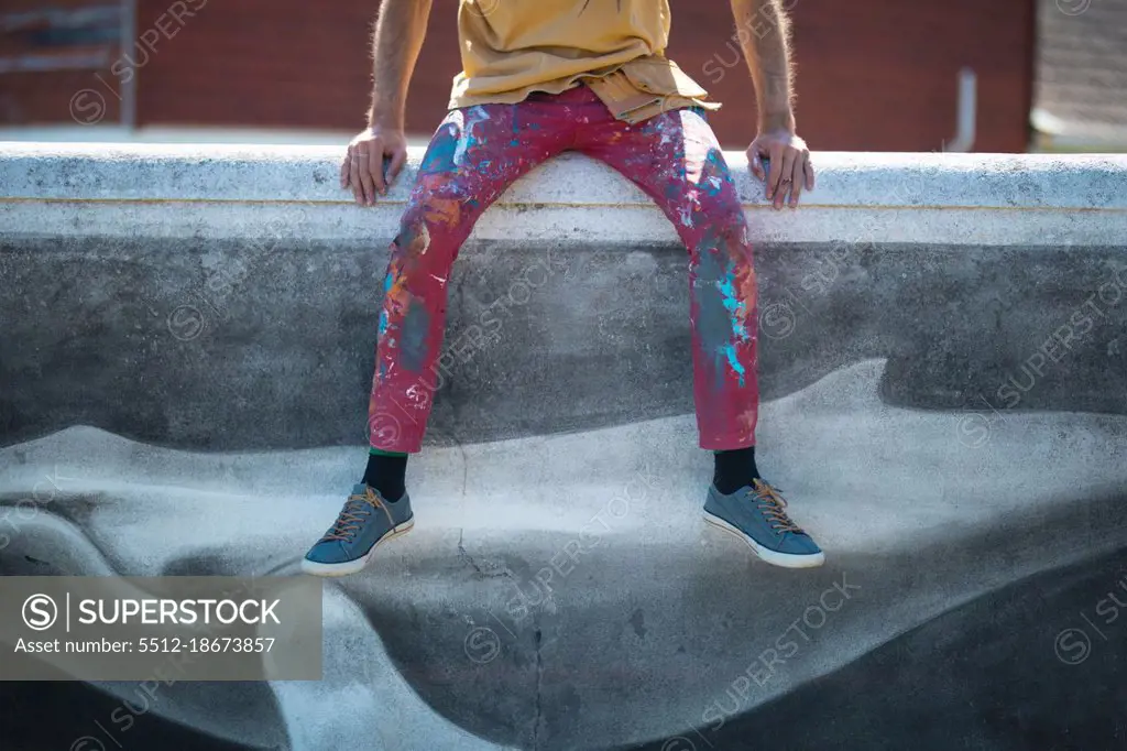 Low section of male artist with messy pant sitting on wall with creative mural painting of wall. street art, creativity and skill.