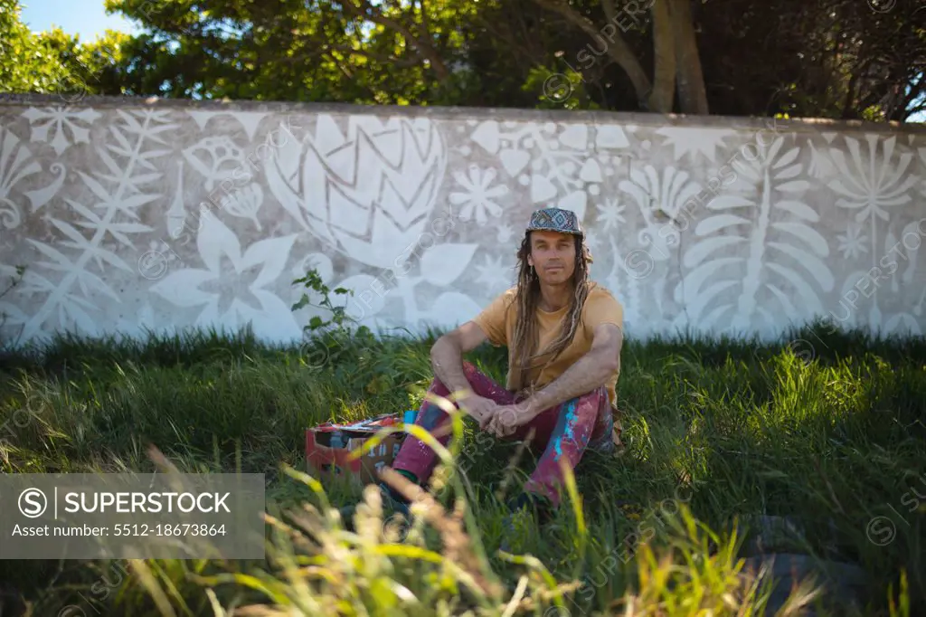 Portrait of male artist sitting on grass against abstract mural painting on wall. street art and skill.