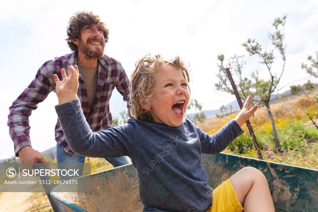 Happy young man pushing excited son shouting while sitting in wheelbarrow at farm. family, homesteading and enjoyment.