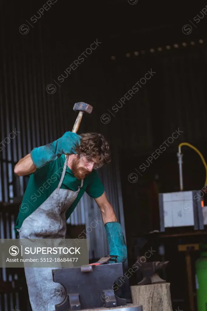 Caucasian bearded blacksmith in protective workwear forging with hammer on anvil in industry. forging, metalwork and manufacturing industry.
