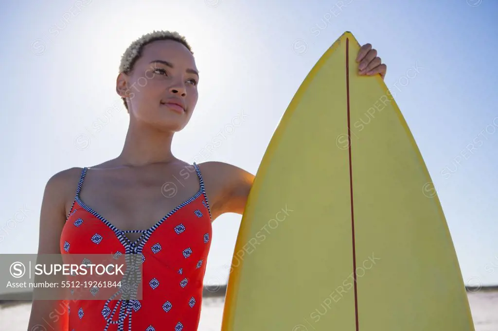 Low angle view of beautiful African american woman in swimwear standing with surfboard on beach in the sunshine