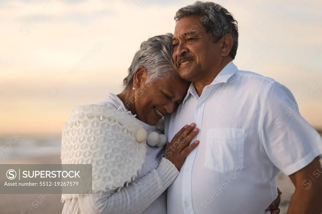 Smiling senior biracial woman embracing man looking away while standing at beach during sunset. lifestyle, love and weekend.