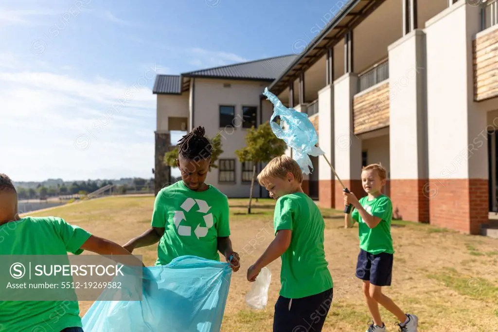 Multiracial elementary schoolboys cleaning ground by school building during sunny day. unaltered, sustainable lifestyle, education, teamwork, cleaning, responsibility and recycling concept.