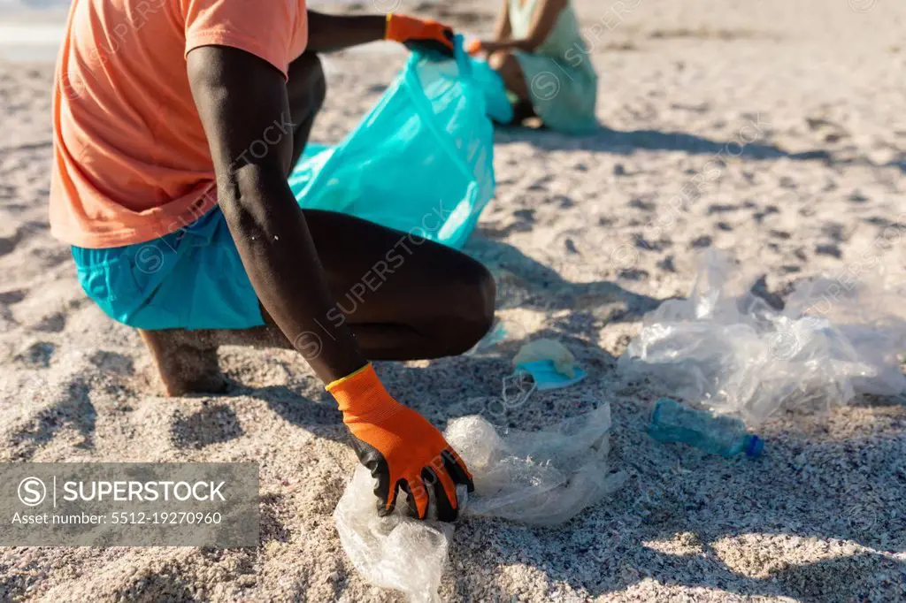 Low section of african american man collecting garbage in plastic bag with girlfriend in background. unaltered, togetherness, responsibility and environmental issues concept.