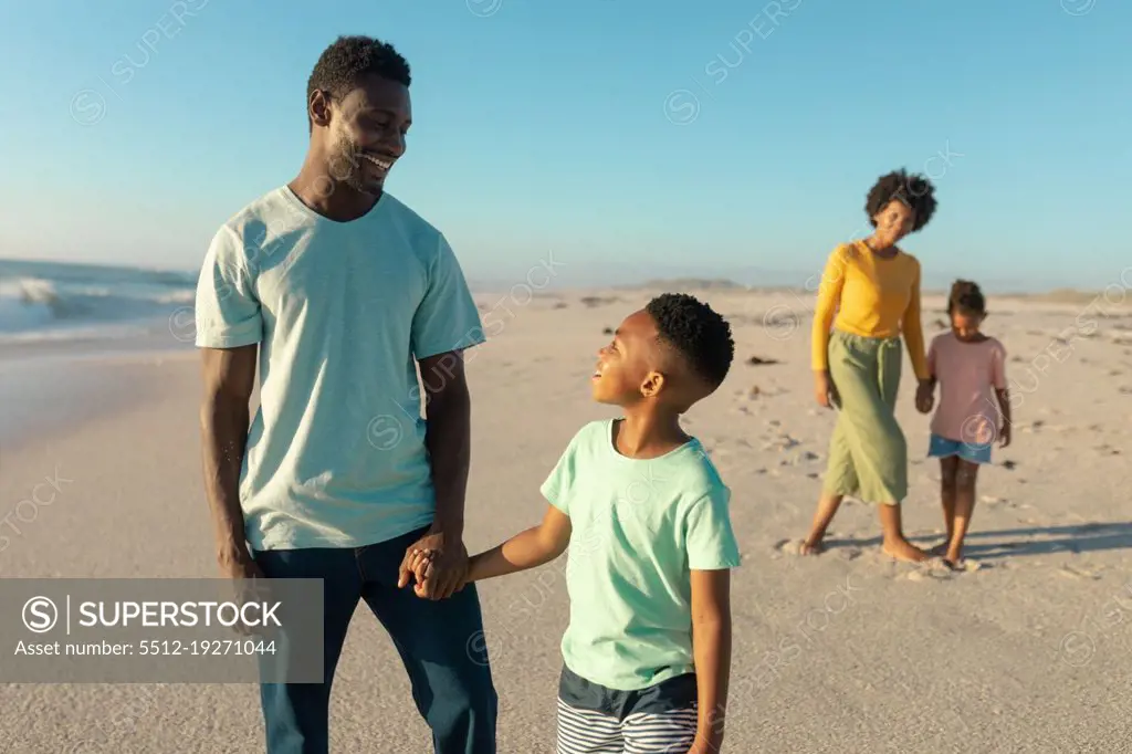 Happy african american father and son holding hands with mother and daughter in background at beach. unaltered, family, lifestyle, togetherness, enjoyment and holiday concept.