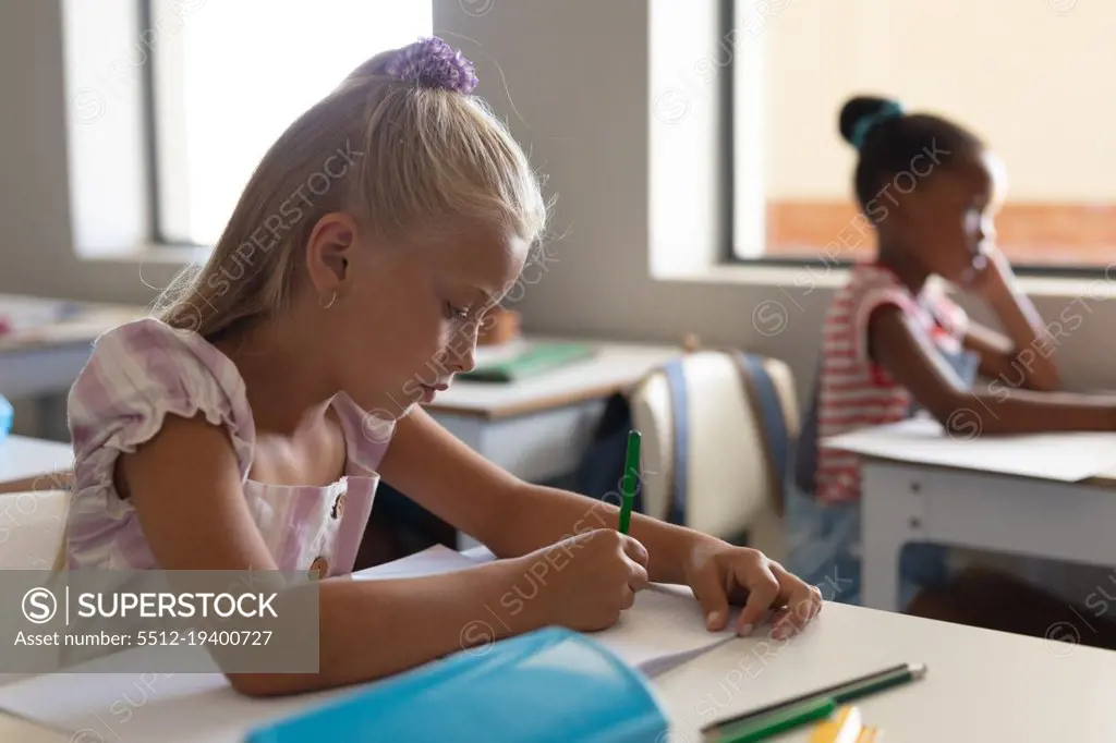 Caucasian elementary schoolgirl writing while studying at desk in classroom. unaltered, teacher, education, learning, studying and school concept.