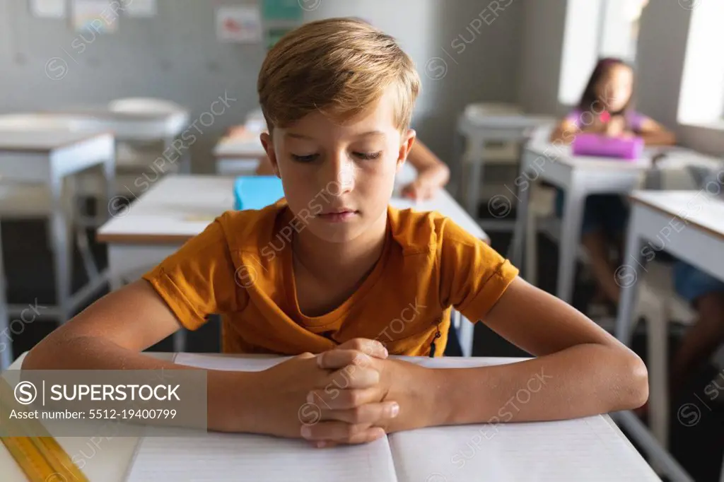 Caucasian elementary schoolboy with hands clasped studying at desk in classroom. unaltered, education, learning, childhood, studying and school concept.