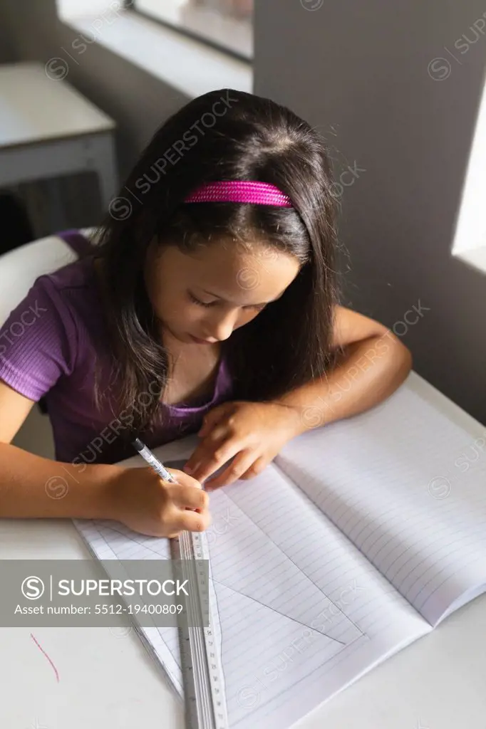 High angle view of caucasian elementary schoolgirl drawing line on book at desk in classroom. unaltered, education, learning, childhood, studying and school concept.