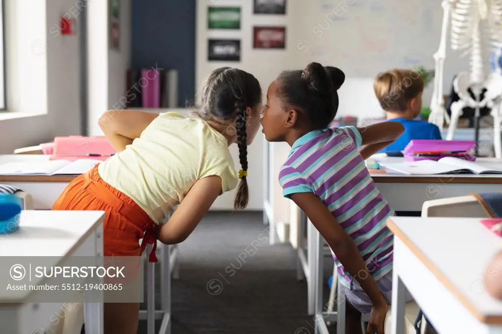 African american elementary girl whispering to caucasian female classmate at desk in classroom. unaltered, education, childhood, gossip, secret, sharing, science, stem and school concept.