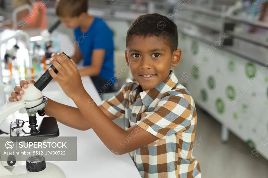 Portrait of biracial elementary schoolboy holding microscope while sitting at desk in laboratory. unaltered, education, childhood, learning, science, stem and school concept.