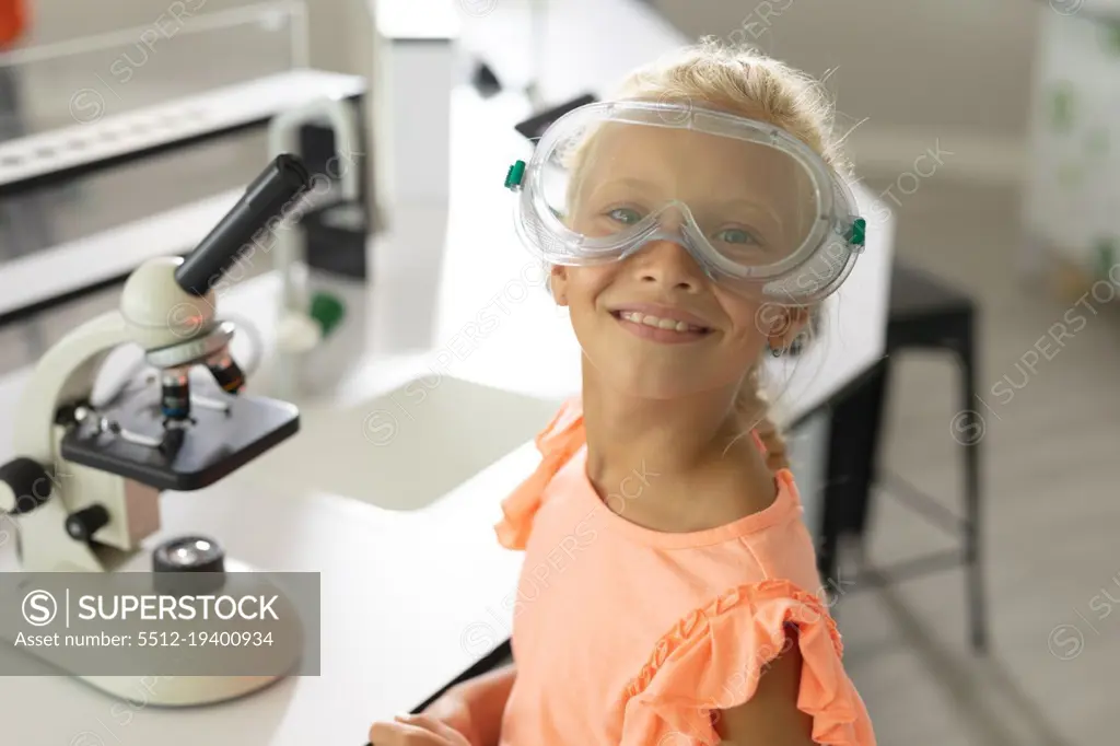 Portrait of smiling caucasian elementary schoolgirl with protective glasses sitting in laboratory. unaltered, education, childhood, learning, science, stem and school concept.