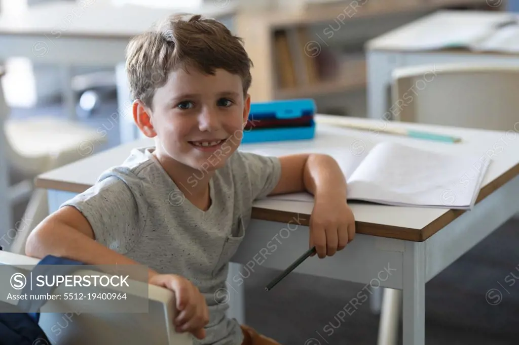 Portrait of cute smiling caucasian elementary schoolboy sitting at desk in classroom. unaltered, education, childhood, studying and school concept.
