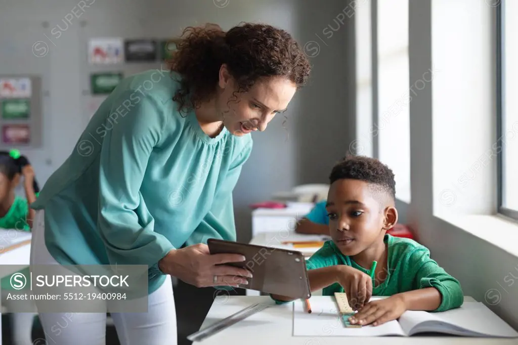 Caucasian young female teacher showing digital tablet to african american elementary boy in class. unaltered, education, childhood, occupation, technology, teaching and school concept.