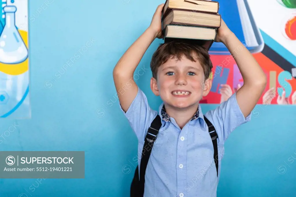 Portrait of smiling caucasian elementary schoolboy stacking books on head against wall in school. unaltered, education, childhood and school concept.