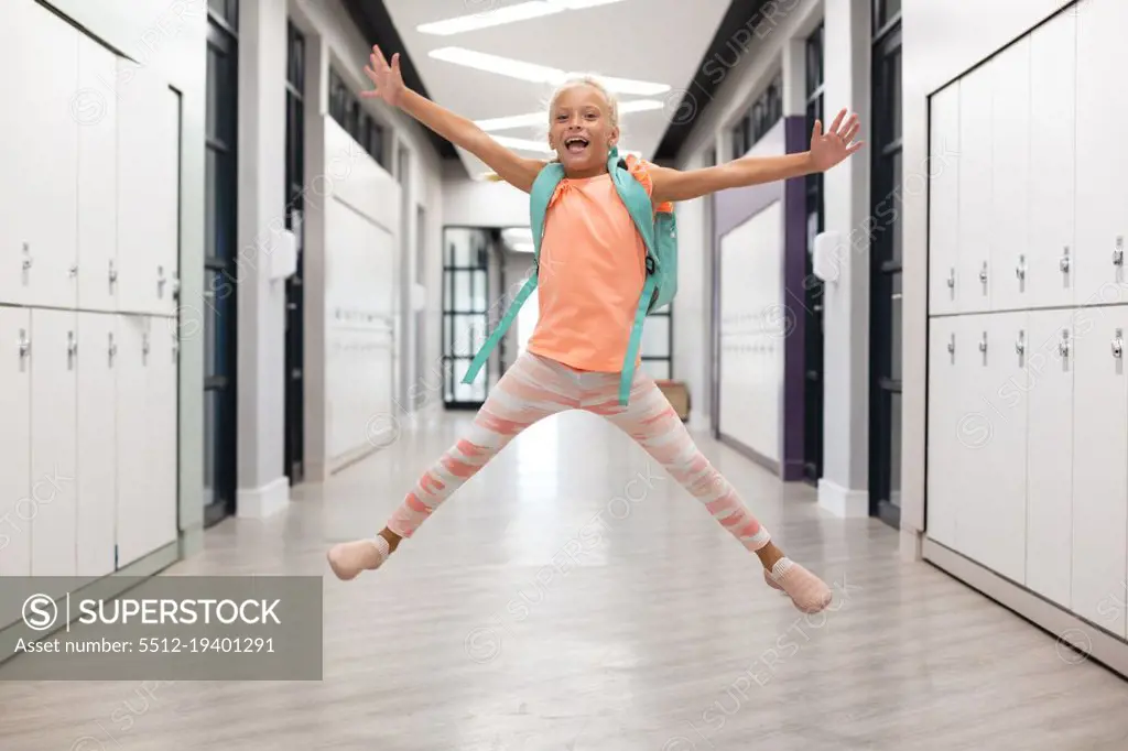 Full length of caucasian elementary schoolgirl with arms outstretched jumping in corridor. unaltered, childhood, education, happiness, playful and school concept.