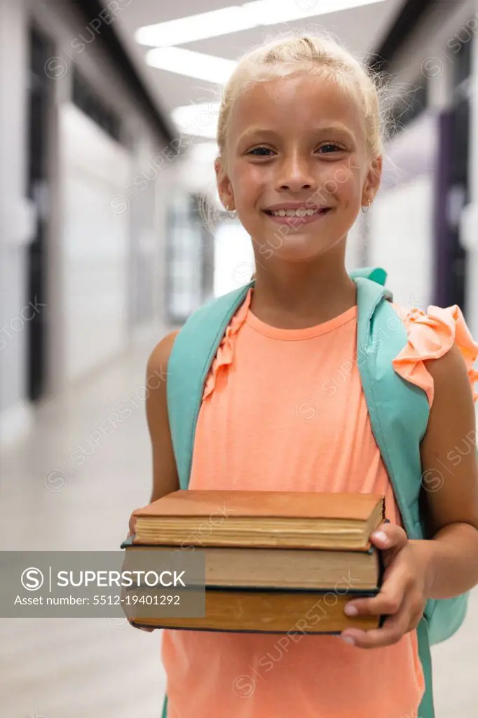 Portrait of smiling caucasian elementary schoolgirl holding books while standing in corridor. unaltered, childhood, education, happiness, learning and school concept.