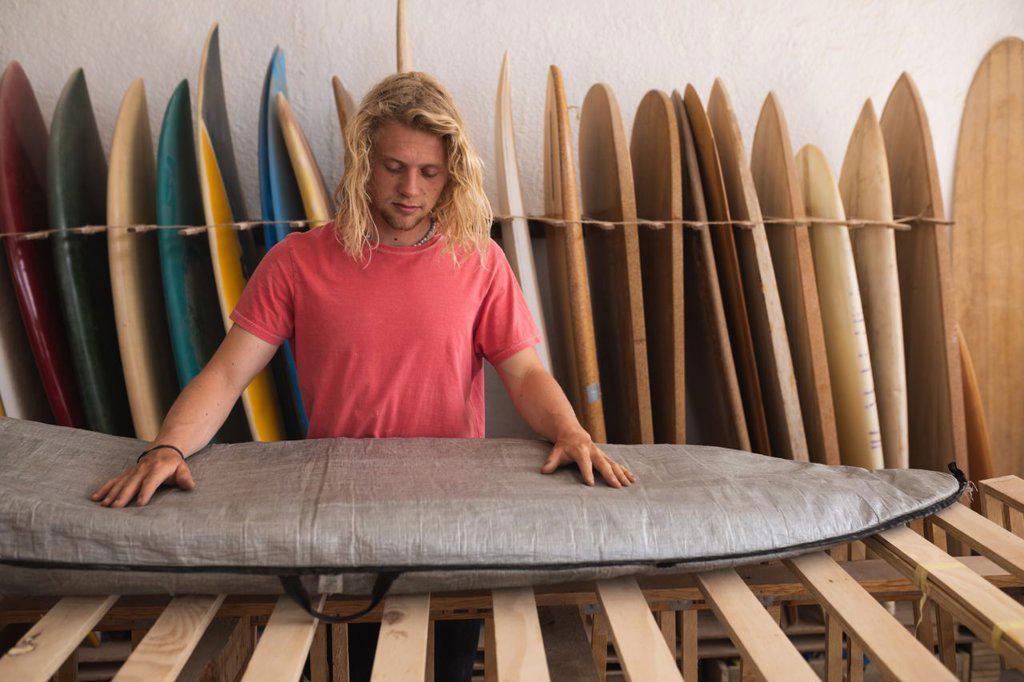 Caucasian male surfboard maker working in his studio, inspecting a surfboard covered with a grey case, with surfboards in a rack in the background.